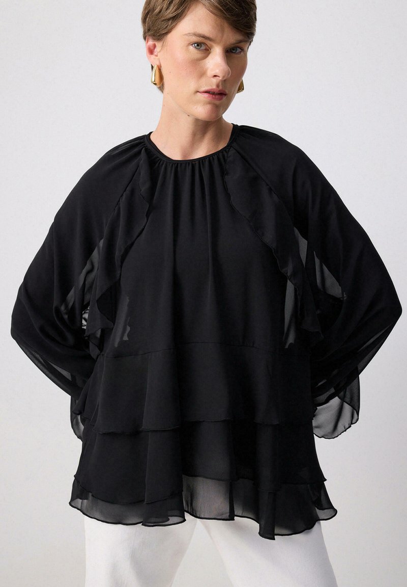 Sort chiffon bluse med lagdelt rynkedesign, brede ærmer og rund halsudskæring. Kombineret med hvide bukser for kontrast.
