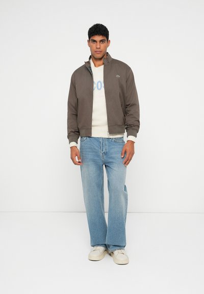 Homme portant une veste zippée marron sur un pull blanc, un jean large bleu clair et des baskets blanches, se tenant devant un fond blanc uni.