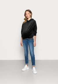 Hoodie de veludo preto com cordão, combinado com jeans azuis ajustados e sapatilhas brancas. O modelo está de pé contra um fundo simples.