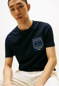 T-shirt in cotone blu navy con un patch logato stampato con la scritta "HILFIGER NEW YORK 1985" in bianco e blu sulla parte sinistra del petto.