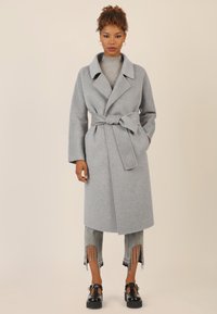 Cappotto di lana grigio con vita cinturata, ampi revers e maniche lunghe, abbinato a jeans grigi scoloriti e scarpe con tacco nero.