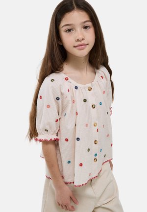 Jeune fille aux longs cheveux bruns portant un chemisier crème avec des fleurs brodées colorées et des boutons en bois, associé à un pantalon clair.