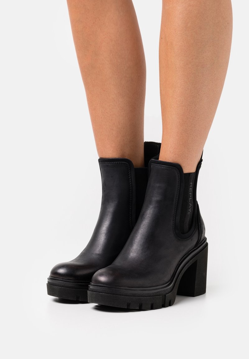 Bottes cheville en cuir noir avec talons carrés épais et panneaux élastiques sur les côtés, portées sur des jambes nues, sur fond blanc.