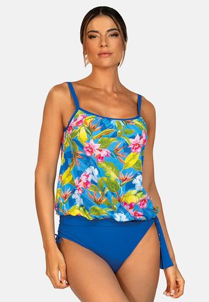Kobieta ubrana w niebieski dwuczęściowy strój kąpielowy z kwiatowym nadrukiem na górze i jednolitym dołem związanym na biodrze, stojąca na jednolitym tle.