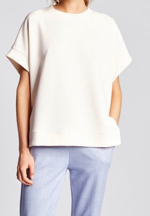 Vrouw die een los wit shirt met korte mouwen en omgeslagen manchetten draagt, gecombineerd met lichtblauwe broek, staat met één hand in haar zak.