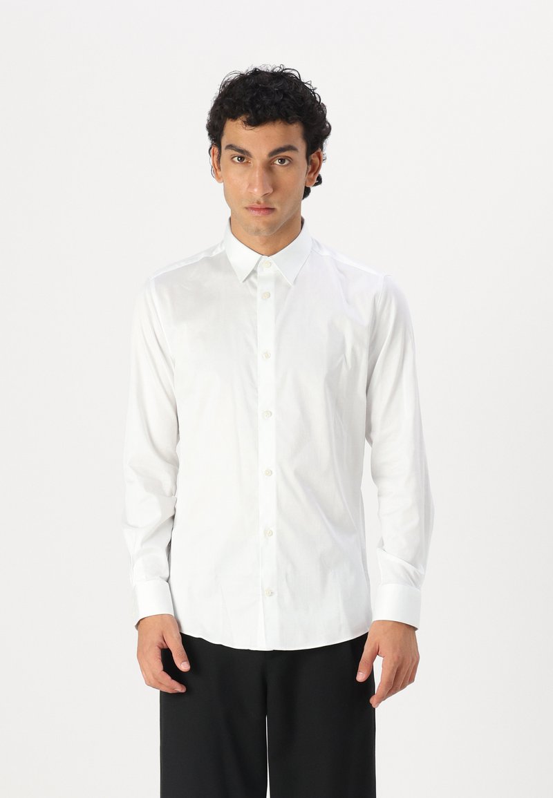 Camisa blanca de manga larga elaborada en un tejido suave, con cuello abotonado y dobladillo recto. Se lleva con pantalones negros.