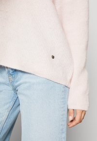 Lichtroze gebreide trui met een ribbeltextuur en een klein metallic logoaccent. Gecombineerd met lichtblauwe denim jeans.