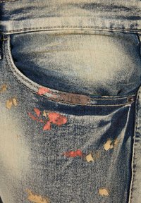 Blå slitte denimjeans med malingssprut i røde og beige toner, med en sølvfarget knapp og forsterkede stikninger i lommene.