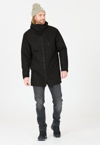 Giacca nera impermeabile con zip frontale, cappuccio regolabile e tasche. Indossata con jeans grigi e scarponi da trekking neri. Sfondo neutro.