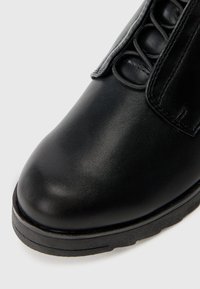Bottine en cuir noir à bout arrondi, surface texturée et accents élastiques sur le côté. Semelle en caoutchouc épaisse pour la durabilité.