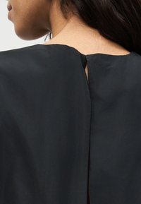 Κοντινό πλάνο λαιμού και ώμου ενός ατόμου που φοράει μαύρο ένδυμα με μικρή σχισμή στην πλάτη στο ύψος του λαιμού, με σκούρα μαλλιά να πέφτουν πάνω στον ώμο.