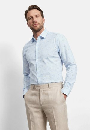Man wearing light blue patterned long-sleeve shirt tucked into beige linen trousers, standing with hands in pockets.