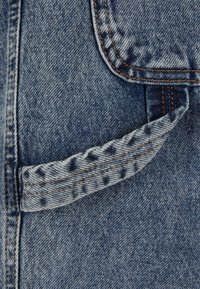 Nära bild av denimtyg som visar en ficka med mönstrad trim och orange sömmar. Texturen är slät med en klassisk blå färg.