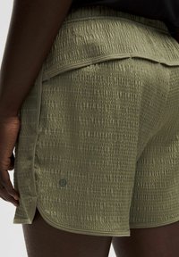 Olīvzaļie teksturētie šorti ar savilktu audumu, elastīgu jostasvietu un sānu slīpēm. Uz apakšas ir neliels logotips.