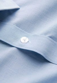 Tissu de chemise en coton bleu clair avec bouton blanc et texture à fine grille, montrant un ourlet cousu et une partie de col.