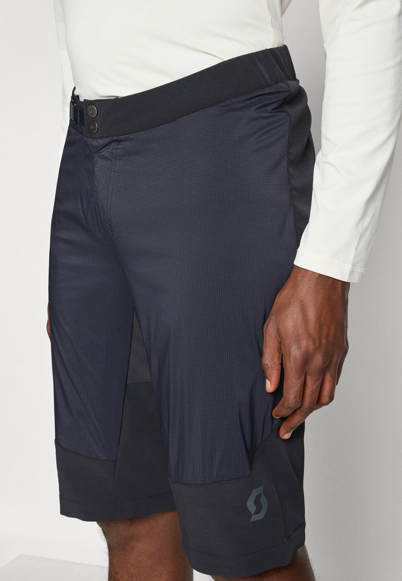 Mann trägt schwarze und dunkelblaue Sportshorts mit einem Doppelknopf am Bund und einem kleinen Logo nahe dem linken Saum.