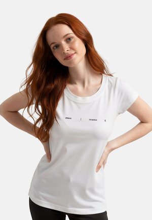 Donna con lunghi capelli rossi che indossa una t-shirt bianca Armani Exchange e pantaloni neri, in posa con le mani sui fianchi su uno sfondo semplice.
