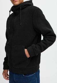 Black fleece hoodie with a front zipper, adjustable drawstring hood, and two side pockets. Soft texture and elastic cuffs and hem.