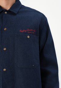 Tmavomodrá denimová košeľa s gombíkovým zapínaním, s náprsníkovou vreckom a vyšitým nápisom "Replay Denim Co. Since 1981" v červenej farbe.