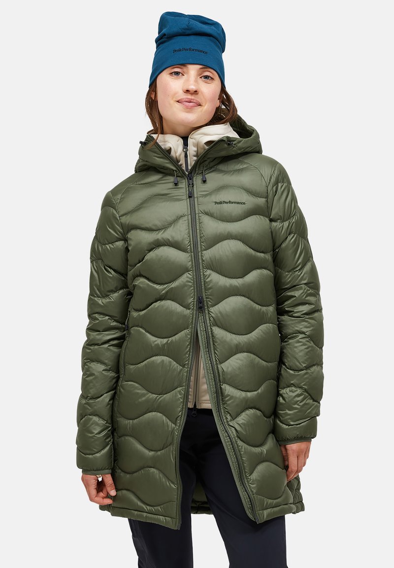 Zöld puff coat hullámos mintával, cipzáras záródással és állítható kapucnival. Egy világos bézs felső fölé van húzva, kék sapkával kiegészítve.