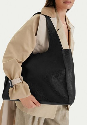 Žena v béžovém trenčkotu držící přes rameno velkou černou koženou kabelku s texturou na bílém pozadí.