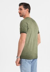 Olivově zelené tričko s krátkým rukávem a texturovaným vzhledem, kulatým výstřihem a ohrnutými manžetami, v kombinaci se světle modrými džínami.