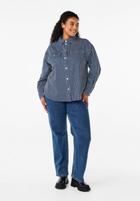 Camicia con bottoni a righe blu e bianche, con colletto e due tasche sul petto. Abbinata a jeans in denim blu e scarpe nere.