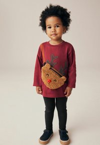 Chemise à manches longues bordeaux avec un motif de bois de cerf, leggings noirs et baskets bleu marine ; sac à épaule en peluche marron en forme de renne avec un nez rouge.