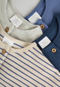 Drei Babyoberteile: hellgrau, marineblau und gestreift in Beige mit blauen Linien. Sie haben runde Ausschnitte und Holzknöpfe. Etiketten sind sichtbar.
