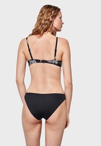 Bikini negro con una tira trasera estampada que presenta detalles florales; parte inferior negra sólida con una textura suave; diseño de cobertura mínima.