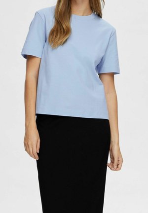 Personne portant une chemise à manches courtes bleu clair rentrée dans une jupe noire taille haute sur un fond uni.