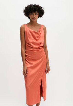 Vrouw met krullend haar die een mouwloze koraalkleurige satijnen jurk draagt met een watervalhals en een voorste split, staand tegen een effen achtergrond.