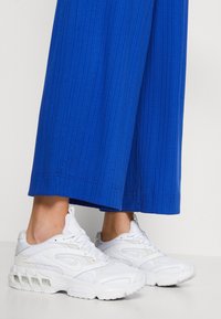 Baskets blanches avec un dessus en maille et cuir, présentant un design distinctif, une semelle rembourrée et une unité d'air proéminente, portées avec un pantalon côtelé bleu.