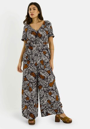Femme portant une combinaison pantalon à jambes larges avec imprimé feuilles noir, blanc et marron, col en V, manches courtes et sandales à plateforme à talons blocs.
