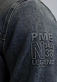 Veste en denim foncé avec un logo brodé distinctif sur l'épaule, mettant en avant "PME N 34°21 38° LEGEND" en fil blanc texturé.
