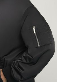 Chaqueta bomber negra con acabado satinado. Presenta un bolsillo con cremallera en la manga, puños acanalados y un diseño estructurado con una textura suave.