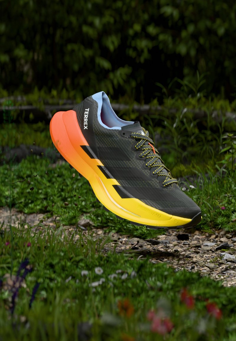 Αθλητικό παπούτσι σε μαύρο και κίτρινο με υφή στην επάνω επιφάνεια, αιωρούμενο πάνω από το χορτάρι. Διαθέτει φωτεινά κορδόνια, μαλακό πάτο και σήμανση "TERREX".