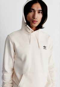 Felpa crema in misto cotone, con tasca a canguro, logo nero di adidas sul petto e cappuccio con cordino. Texture liscia.