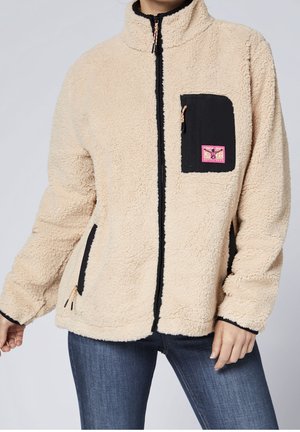 Beżowa kurtka z polaru z czarnym zamkiem błyskawicznym i kieszeniami, różowa naszywka na kieszeni na piersi, noszona z niebieskimi dżinsami na osobie stojącej na jednolitym tle.