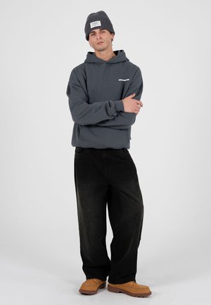 Grauer Hoodie mit kleinem weißem Text, schwarze Jeans mit weitem Bein und beige Slip-On-Schuhe. Das Model trägt eine graue Strickmütze. Einfarbiger Hintergrund.