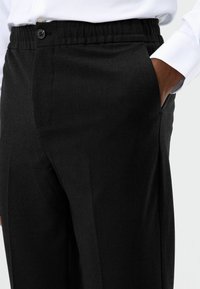 Zwarte broek met een gestructureerde stof. Voorzien van een elastische tailleband met een knoop en zijzakken. Strakke, op maat gemaakte snit.