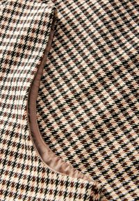 Tkanina o wzorze hounds tooth w kolorze beżowym, czarnym i rdzawym, o gładkiej teksturze oraz kontrastową podszewką widoczną wzdłuż krawędzi.