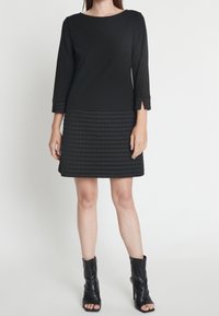 Svart klänning med en åtsittande överdel och en texturerad, vadderad kjol. Trekvartslånga ärmar har vikta detaljer. Bärs med svarta ankelstövlar.