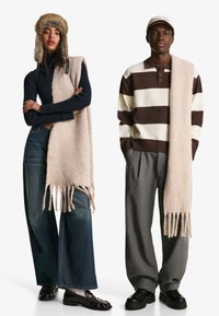 Due individui indossano abiti invernali. Il primo ha un dolcevita a coste blu navy e una sciarpa frangiata marrone chiaro. Il secondo indossa un maglione a righe marrone e crema con una sciarpa leggera. Entrambi hanno pantaloni larghi e calzature scure.