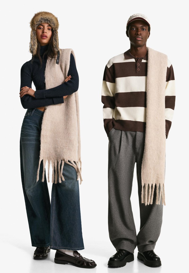 Due individui indossano abiti invernali. Il primo ha un dolcevita a coste blu navy e una sciarpa frangiata marrone chiaro. Il secondo indossa un maglione a righe marrone e crema con una sciarpa leggera. Entrambi hanno pantaloni larghi e calzature scure.