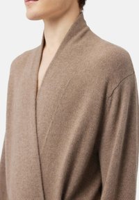 Brun, mjukstickad cardigan med sjalkrage och avslappnad passform. Har långa ärmar och en minimalistisk design som betonar textur och värme.