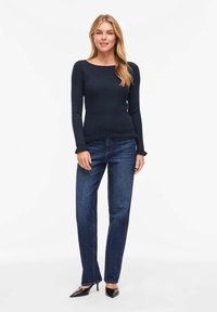 Femme debout portant un haut à manches longues côtelé sombre, un jean bleu droit et des talons noirs à bout pointu, sur un fond blanc.