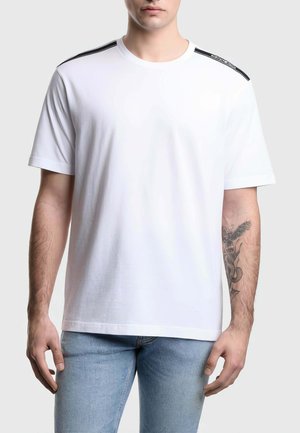 Uomo che indossa una maglietta bianca semplice a maniche corte con una striscia nera sulla spalla e jeans blu, mostrando un tatuaggio sull'avambraccio sinistro.