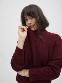 Suéter de malha bordô com gola alta e punhos canelados. Tecido texturizado com corte amplo e ombros caídos. Modelo posa com a mão perto da gola.