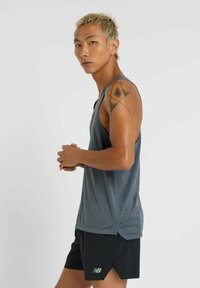 Grijze sportieve tanktop met een gestructureerd oppervlak, gecombineerd met zwarte shorts met zijsplitten en een wit logo. Model poseert zijwaarts.